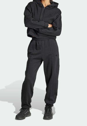 Osoba ubrana w czarną bluzę Adidas na zamek, dopasowane czarne spodnie dresowe i czarne sneakersy, stojąca z założonymi rękami.