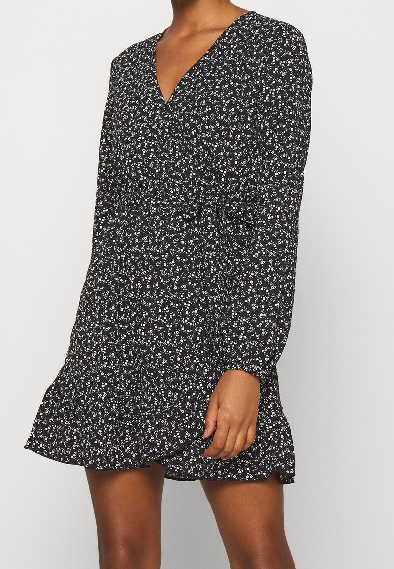 Femme portant une robe portefeuille noire à manches longues avec imprimé floral blanc, encolure en V, et ourlet à volants, debout sur un fond uni.