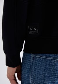 Černá mikina s texturovaným povrchem, která má obdélníkovou značkovou etiketu s logem "A|X" a textem "Armani Exchange Blue Edition".