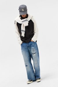 Άτομο που φοράει μαύρο και κρεμ φούτερ, λευκό κασκόλ με σχέδια, σκισμένα μπλε τζιν, αθλητικά παπούτσια και καπέλο με σχέδια, στέκεται με τα χέρια στις τσέπες.