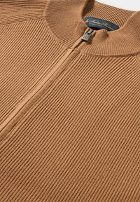 Nahaufnahme eines beigefarbenen, gerippten Strickpullovers mit Reißverschluss und einem marineblauen Etikett im Kragen.