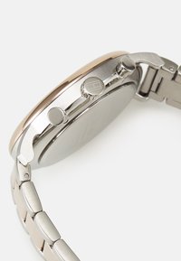Reloj de acero inoxidable con un bisel en oro rosa, caja redonda y pulsera de eslabones metálicos. Cuenta con dos botones en el lateral y superficies pulidas.