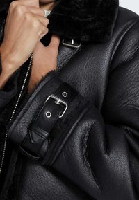 Personne en train de boucler le col d'une veste en cuir noir avec une finition texturée et des sangles à boucle au niveau des manches et du col.