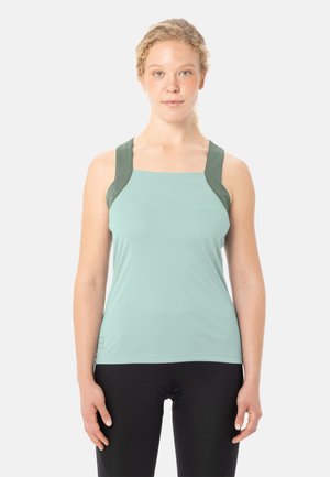 Femme à la peau claire et aux cheveux blonds bouclés portant un haut de sport sans manches vert clair et un legging noir, debout devant un fond blanc.