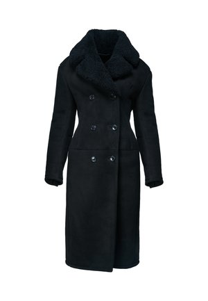 Femme portant un long manteau noir croisé avec un grand col, des bottes noires montantes, tenant un sac à main noir, debout devant un fond uni.