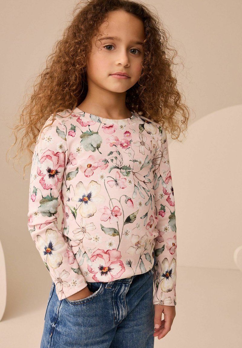 Enfant aux cheveux bouclés portant un haut à fleurs à manches longues et un jean bleu, debout avec une main dans la poche devant un fond neutre.