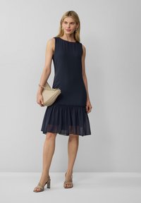 Navy mouwloze jurk met een gerimpelde zoom; gemaakt van een gladde stof. Gepaard met een beige handtas en sandalen met blokhak en banden.
