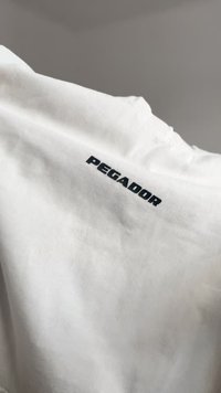 Λευκό βαμβακερό μπλουζάκι με την ένδειξη "PEGADOR" σε έντονα μαύρα γράμματα στην πλάτη. Το ύφασμα έχει απαλή υφή με απλό σχέδιο.