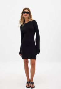Juoda juostos suknelė su ilgomis rankovėmis, prigludusia siluetu, apvaliu iškirpimu ir virš kelių ilgio. Modelis dėvi juodus akinius nuo saulės ir sandalus.
