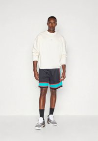 Crèmekleurige sweatshirt met geribbelde manchetten, gecombineerd met zwarte korte broek met turquoise en rode strepen. Het model draagt grijze sneakers en zwarte sokken.