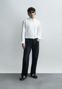 Camisa branca de botões, jeans escuros e sapatos pretos com riscas brancas. A camisa tem mangas compridas e os jeans são de corte direito.