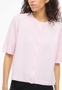 Naine, kes kannab pehmet roosat lühikeste varrukatega nööpidega kardigani ja musti pükse, näidatud õlgadest vööni.