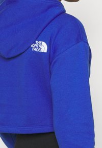 Felpa blu cropped realizzata in un tessuto morbido. Presenta un logo bianco sulla schiena e un cappuccio con cordino. Dettaglio delle cuciture visibile sull'orlo.