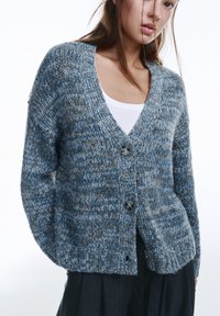 Grob gestrickter Cardigan in Blau mit dezenten braunen Sprenkeln, mit V-Ausschnitt, zwei dunklen Knöpfen und gerippten Bündchen. Weiche Textur und entspannte Passform.