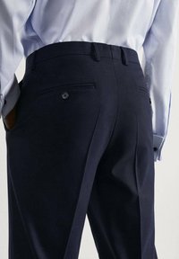 Marineblauwe pantalon met een getailleerde pasvorm, voorzien van een gladde textuur, twee achterzakken en een knoopdetail. G gedragen met een lichtblauw shirt.