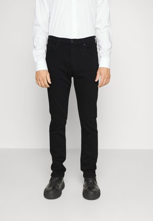 Personne portant un pantalon noir slim, des baskets noires et une chemise blanche à manches longues boutonnée, debout devant un fond uni.