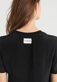 Žena s dlouhými hnědými vlasy, která má na sobě černé tričko s malou bílou nálepkou s nápisem "CALVIN KLEIN JEANS" na horní části zad.