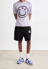 Lavendel grafisk t-shirt med sort cirkulært logo, parret med sorte shorts med et syet logo. Blå og hvide sneakers fuldender outfittet.