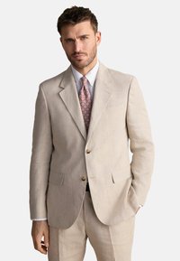 Costume en lin beige avec un blazer à un bouton, revers en pointe et boutons dorés. Associé à une chemise bleu clair et une cravate à motifs roses.