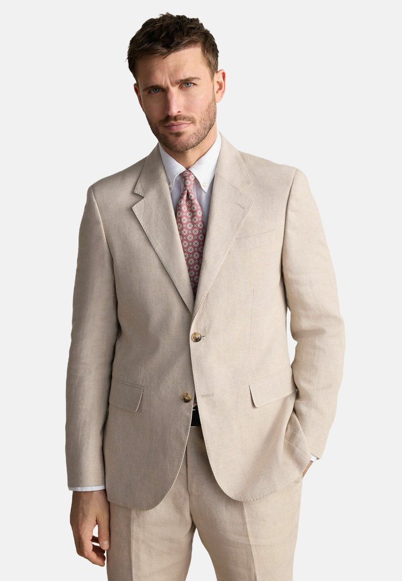 Costume en lin beige avec un blazer à un bouton, revers en pointe et boutons dorés. Associé à une chemise bleu clair et une cravate à motifs roses.