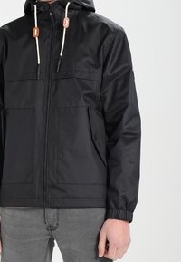 Zwarte capuchonjas met lichte trekkoorden, ritssluiting aan de voorkant, twee borstzakken en elastische manchetten. Gemaakt van waterafstotend materiaal.