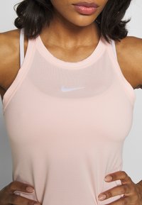 Donna che indossa un top atletico senza maniche Nike di colore rosa chiaro, con le mani sui fianchi, mostrando il busto superiore e le labbra.