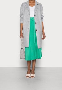Cappotto lungo grigio, top bianco, gonna plissettata verde, décolleté bianchi aperti, e borsetta grigia chiara. Tessuto testurizzato con chiusura a bottone.