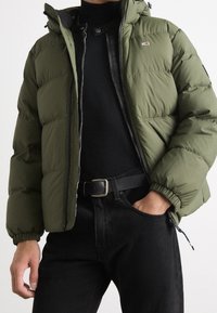 Olivgrön puffjacka med huva, har en frontdragkedja och elastiska ärmslut. Bärs över en svart polotröja och svarta jeans med bälte.