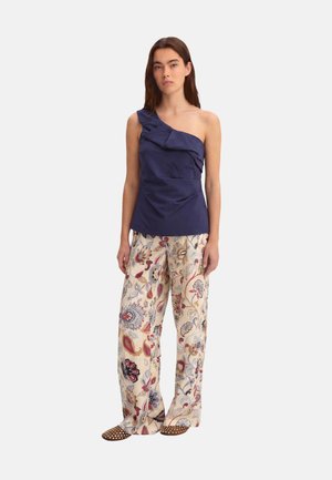 Donna con top blu navy monospalla, pantaloni larghi color crema con motivi floreali colorati e scarpe marroni a pois, in piedi su sfondo bianco.
