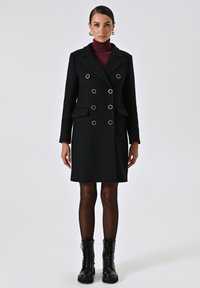 Cappotto nero doppiopetto con hardware dorato, spalle strutturate, due tasche frontali e lunghezza fino a metà coscia. Il modello indossa stivali neri alla caviglia.