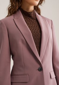 Zeleni blazer vijolične barve s prilagojeno obliko, z režnimi ovratniki in z enim gumbom za zapenjanje. Pod njim je rjav teksturiran zgornji del. Gladek material. Sprednji žep.