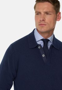 Námořnický svetr s límečkem a zapínáním na dvě knoflíky, vyrobený z texturované látky přes světle modrou košili s vzorem a kravatou.