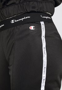 Svarta atletiska byxor med en slät textur, med en midja prydd av logotyp och ett vitt sidostrip som har upprepade logotypdetaljer.