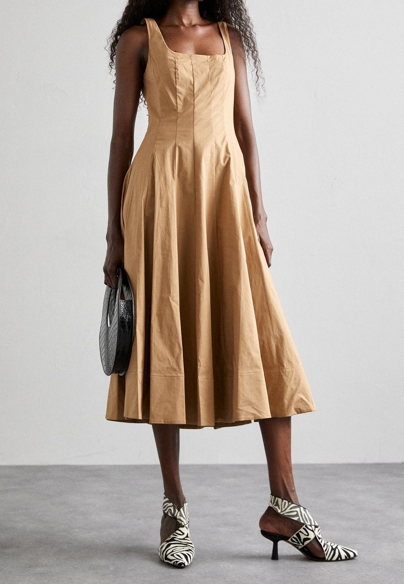 Femme portant une robe midi sans manches beige, tenant un sac à main rond noir et portant des talons hauts à motif zébré sur un sol gris.
