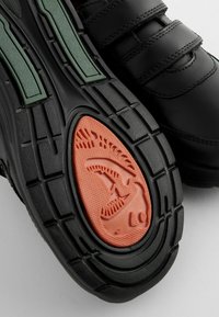 Zapatos negros con acentos verdes, correas de velcro, suela texturizada, con un detalle de goma naranja en forma de tiburón.