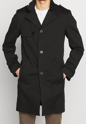 Homme portant un trench-coat noir boutonné, les mains dans les poches, associé à un pantalon marron et une chemise blanche en dessous.