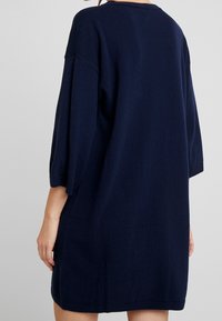 Osoba w luźnej, sięgającej do kolan, ciemnogranatowej sukience sweterowej z rękawami o długości trzech czwartych, pokazana z tyłu.