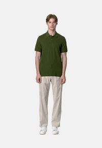 Polo shirt verde oliva con maniche corte e colletto, abbinato a pantaloni beige chiaro e sneakers bianche. Design semplice e aderente.