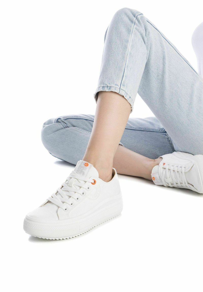 Zapatillas de lona blancas con una parte superior texturizada, punta redonda y suela de goma elevada. Presenta ojales naranjas y un detalle de logo en la lengüeta.