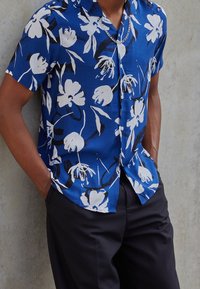Lyhythihainen sininen paita, jossa on valkoisia kukkakuvioita ja mustia yksityiskohtia. Valmistettu kevyestä kankaasta, napitettuna edestä ja rento istuvuus.