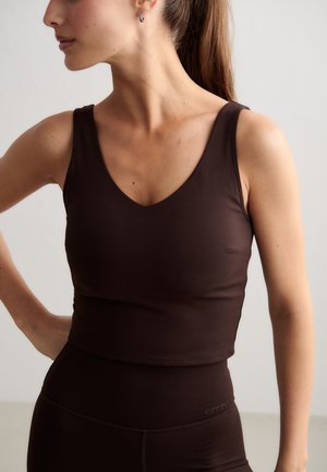 Vrouw met een bruine mouwloze crop top en hoog gerokte legging staat met één hand op de heup en kijkt opzij tegen een effen achtergrond.