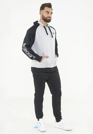Bebaarde man staande met handen in de zakken, draagt een zwart-witte hoodie, zwarte broek en witte sneakers op een witte achtergrond.