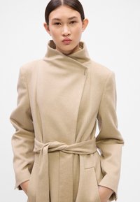 Abrigo de mezcla de lana beige con un cuello alto, diseño envolvente y cintura atada, con bolsillos laterales y un detalle de acento plateado.