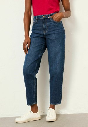 Person wearing high-waist dark blue jeans, red top, and white sneakers standing with one hand in pocket against a plain light wall.