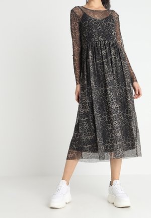 Femme portant une robe midi à manches longues en voile noir à imprimé léopard, superposée sur une combinaison noire, associée à des baskets blanches épaisses.