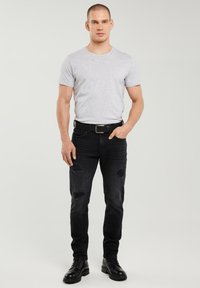 Mand stående iført en lysegrå T-shirt med korte ærmer, sorte revnede jeans, sort bælte og sorte snørebåndsstøvler foran en ensfarvet baggrund.