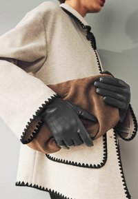Black leather gloves hold a brown fabric clutch. A pale gray coat with a scalloped black trim is visible, featuring a round button closure.