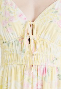 Gulblommig klänning med djup V-ringning, justerbar knytning och elastisk midja. Tyget har en mjuk, lätt textur med rosa och gröna accenter.