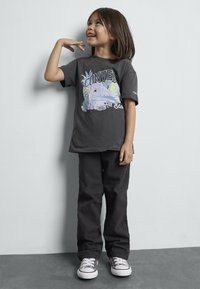 T-shirt gris avec un design graphique coloré représentant des fruits et une sneaker, associé à un pantalon noir ample et des sneakers argentés.
