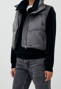 Persona che indossa una felpa nera, un gilet imbottito grigio con tasche con cerniera e jeans neri slavati, in piedi contro uno sfondo chiaro e uniforme.